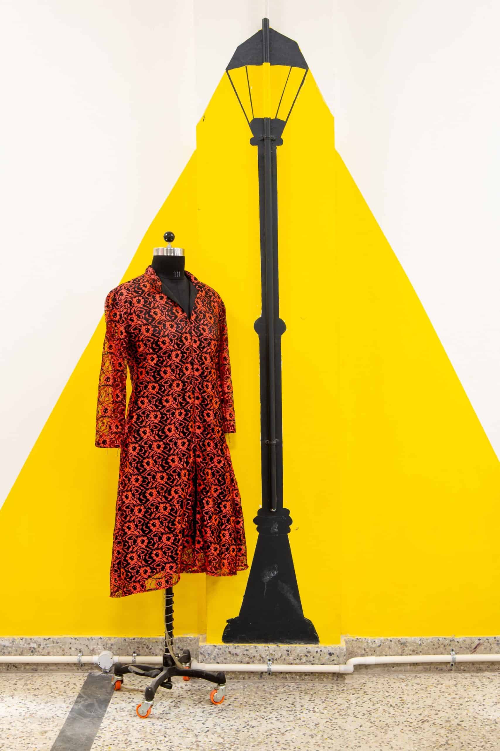 A mannequin dressed in a bright red costume, placed before a yellow and white background beside a lamppost painting